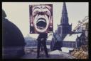 Anonymous, ‘Colour photograph of Dougie Thomson standing on top of a tomb in the Glasgow Necropolis, holding a large clown placard ’ [c.1978]
