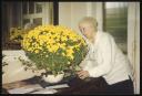 collection owner: Marie-Louise Von Motesiczky, ‘Photograph of Marie-Louise von Motesiczky standing next to a table with a flower-pot holding a plant with lots of yellow flowers ’ [c.1990s]