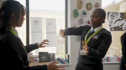 Two students face each other in an art classroom, mirroring each others hand movements and gestures. Student artwork is displayed on the walls and windows behind them