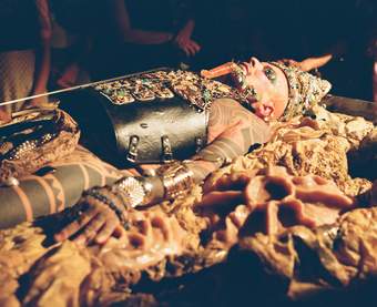 Ron Athey, a white man lies in a bed of silicone viscera. He wears a breast plate and crown and is covered in jewels. His many tattoos are visible.