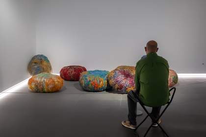 A photograph of a man in a green top sitting on a portable stool in front of an art installation.