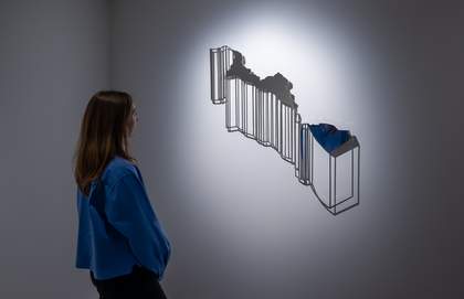 A photograph of a woman in a blue jacket looking at a silver artwork on the wall.