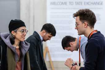 Two people wearing Tate staff lanyards greet guests - a printed sign behind reads 'welcome to Tate Exchange'