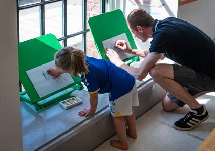 A child and parent crouch down near a window drawing at an easel