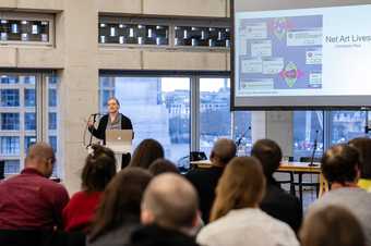 A person stands at a lectern in the Tate Exchange space at Tate Modern, with a projected slide presentation nearby, speaking to an audience