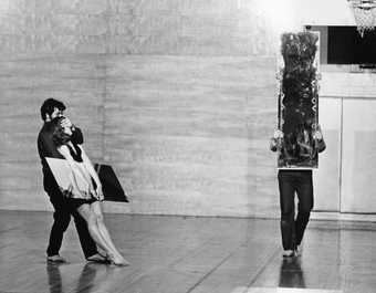 Three performers in an otherwise empty space, one holding a mirror up to the camera, another holding a mirror and being held by the third performer