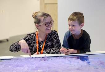 A Tate member of staff is pointing at a 3D map and smiling at a little boy who is standing next to her.