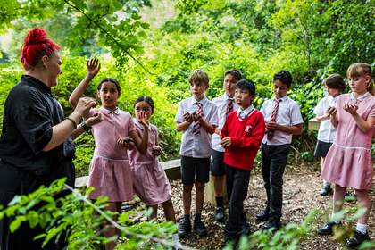 Teacher Lyndsey Miles stands with students in a forest clearing. The students are holding clay sculptures in their hands. The student nearest Lyndsey has their hand raised to ask a question.