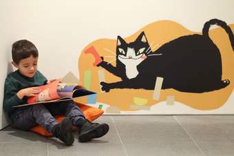 A photograph of a child sat reading a book with a cartoon drawing of Mildred the Gallery Cat on the wall behind