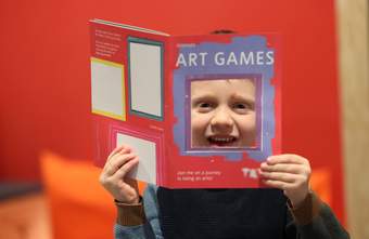 A photograph of a child looking through a square cut out of a booklet that has the words 'Art Games' on the front cover.