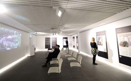 A wide photograph of an exhibition at Tate Liverpool + RIBA North with visitors looking at the photographs on the wall and watching the film