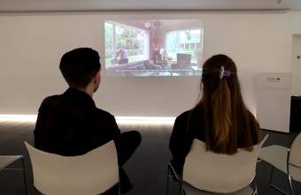 A photograph of two visitors sat watching a film in Tate Liverpool + RIBA North