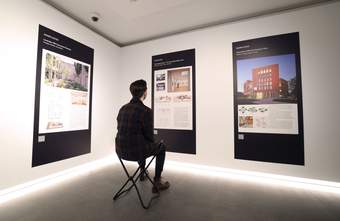 A photograph of a visitor sat on a folding stool whilst looking at a big photo on the wall
