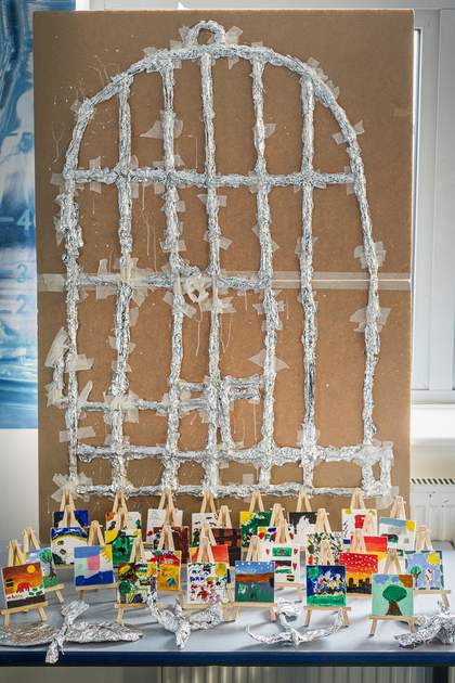 A display of the students’ work. In the background is a large cage structure made of of straws and tinfoil, attached to a large piece of cardboard. Tiny painted canvases sit on easels in the middle, and at the front are four tinfoil birds.