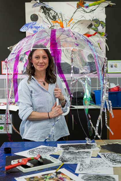 Teacher Clare Boreham Kerr stands in her classroom with mono-prints in front of her and art behind her. She holds one of her sensory umbrellas over her head, and the iridescent and purple ribbons hang down around her