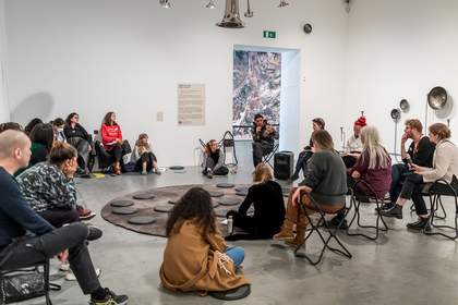 A group of people sit in a gallery space, immersed in discussion.