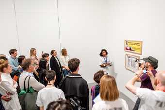 a woman stands in front of a Guerrilla Girls artwork and speaks in front of a crowd in Tate Modern gallery