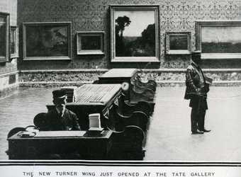 Photo taken of the Turner wing when it has recently opened, with a man looking at paintings on the wall, a warden at the forefront of the photo and tables in the middle of the room.