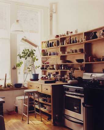 This is an image of a kitchen with wooden shelves full of ceramic vases and pots. There is also a plant, a cooker and a wooden stool in shot.