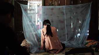 still from a video artwork, showing a person sat in a dark room in front of a thin curtain of fabric