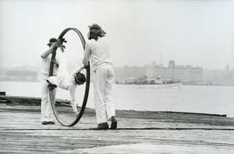 Two performers dressed in white push a third performer held in a large hoop along the side of a river