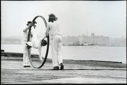 Two figure in white push a large hoop containing another figure in white