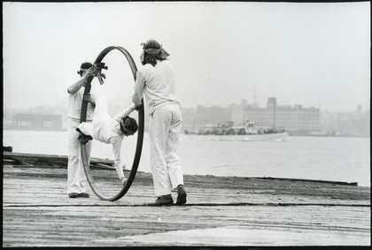 Two figure in white push a large hoop containing another figure in white