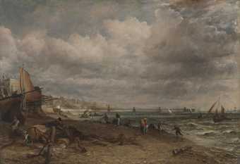 Painting of a beach scene under a grey sky, with Brighton visible along the coast. There are boats of all sizes on the sea and hauled up on the beach, and figures appear on the beach, concentrated at the water’s edge.