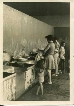 Two women stand in a worn-out space washing clothes at a bank of sinks, with two children nearby