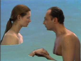 A topless man and woman face each other; behind them, a blue-green sea
