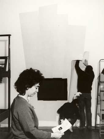 Black and white photo, a person in the foreground looks down at a piece of paper, two people in the background are adhering paper to a gallery wall