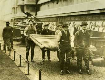 Photo of workmen carrying a large artwork, the exterior of Tate Britain and a vehicle are visible in the background.
