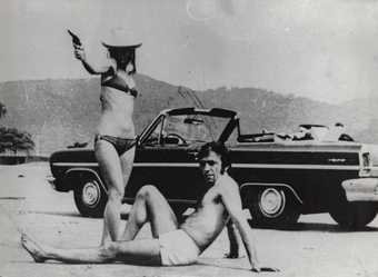 two people on a beach sit or stand with a gun next to a car