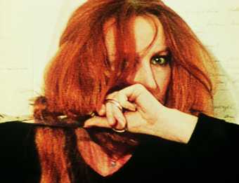 A woman with vibrant ginger hair looks into the camera whilst she appear the cut her hair with scissors.