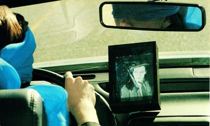 This film still is shot from the backseat of a car. The driver in the front seat wheres a blue blind fold. There is a framed picture of a person to the right of the steering wheel.