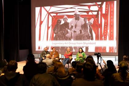 A group of four people are sitting on a stage facing a crowd with an image projected on the wall behind them.