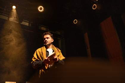 A man in a yellow shawl stands holding a book with spotlights behind him