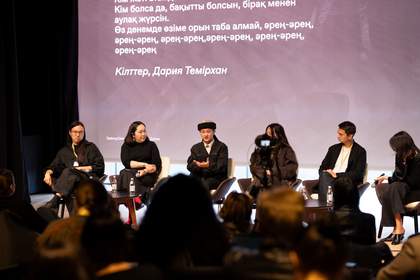 A group of six people sit on a stage with a crowd in the foreground and a projector presentation in the background.