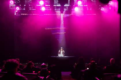 A woman sits by a desk on a stage in front of a group of people on either side.