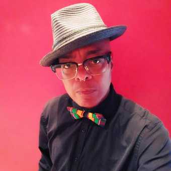 Portrait of Campbell X in front of a pink background, wearing a black shirt, colourful bow tie and hat.