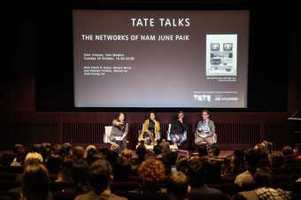 Photograph of Sook-Kyung Lee, Cécile B. Evans, Haroon Mirza and Stephen Vitiello sitting together onstage at Tate Modern. Sook-Kyung Lee reaches out with her hand to take a question from the audience.