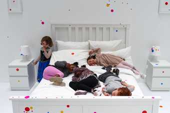 Children playing with colourful dot shaped stickers on a large bed in a white room.