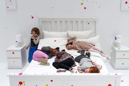 An all white bedroom with a children playing on the bed and a few coloured circles on the wall.