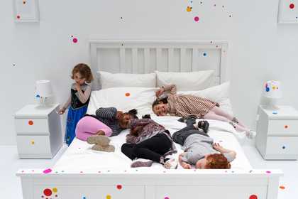 An all white bedroom with a children playing on the bed and a few coloured circles on the wall.