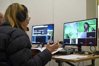 Photograph of the back of a person with headphones on looking at two computer screens of live filming