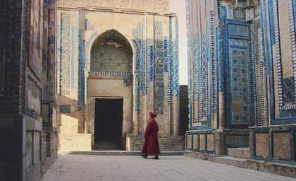 This film still shows a man in maroon robes and turban walking between tall tiled buildings with intricate blue illustrations on each tile.