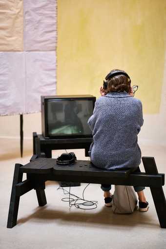 visitor sat on a bench wearing headphones and watching artwork on a tv screen