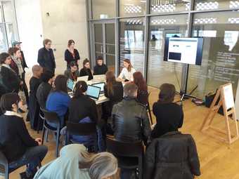 A group of people gather around a table, some of using laptops, listening to a workshop leader, behind whom is a screen displaying their own laptop screen for the audience