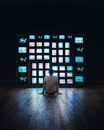 person sat in dark room on floor in front of wall of tv sets