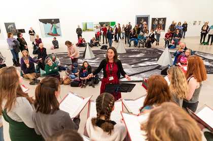 In the foreground 7 young people sing in a choir, with open songbooks and a conductor. There is a gallery installation of a black and white carpet with crystal shapes and cones. People stand and sit listening to the choir.